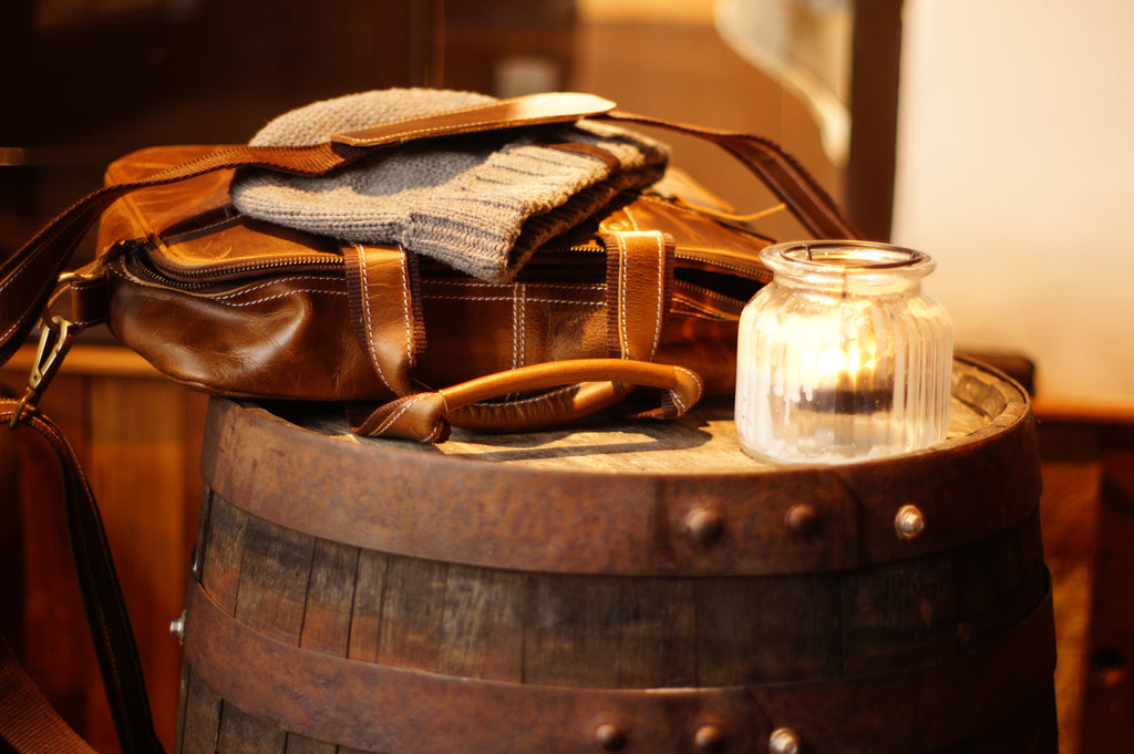 Men's brown leather messenger bag on whiskey barrel with woolie beanie and candle.